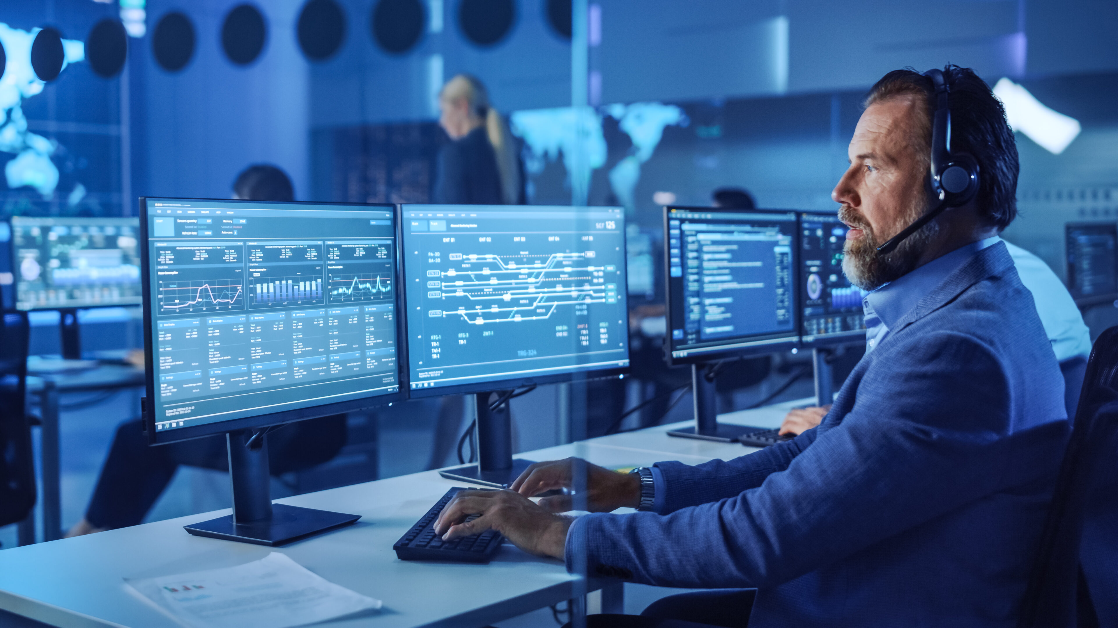 Confident Male Data Scientist Works on Personal Computer Wearing a Headset in Big Infrastructure Control and Monitoring Room. Senior Engineer in a Call Center Office Room with Colleagues.